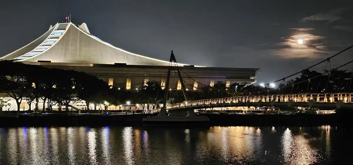 A nighttime photo of a modern building with a distinctive sloped roof, illuminated against a dark sky with a visible moon. A…