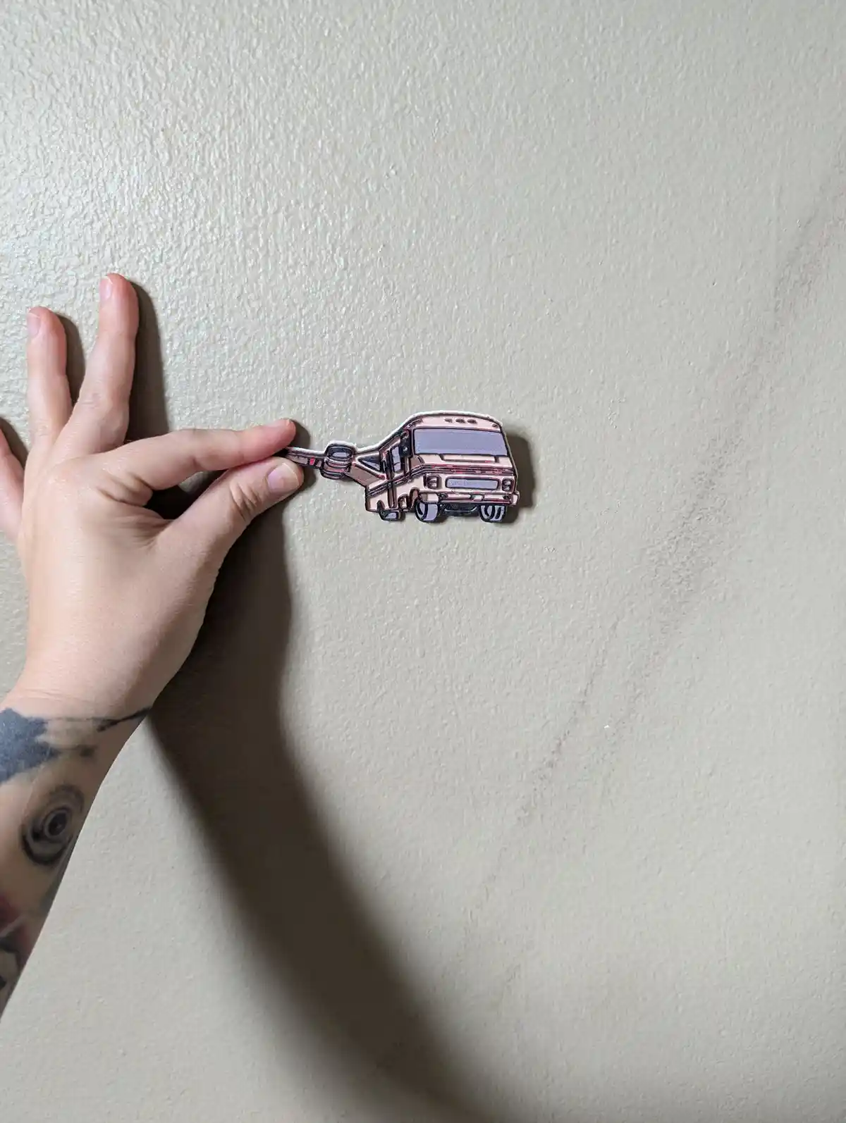 A hand with tattoos holds a small, pinkish RV-shaped magnet against a beige textured wall. The magnet is shaped like a…