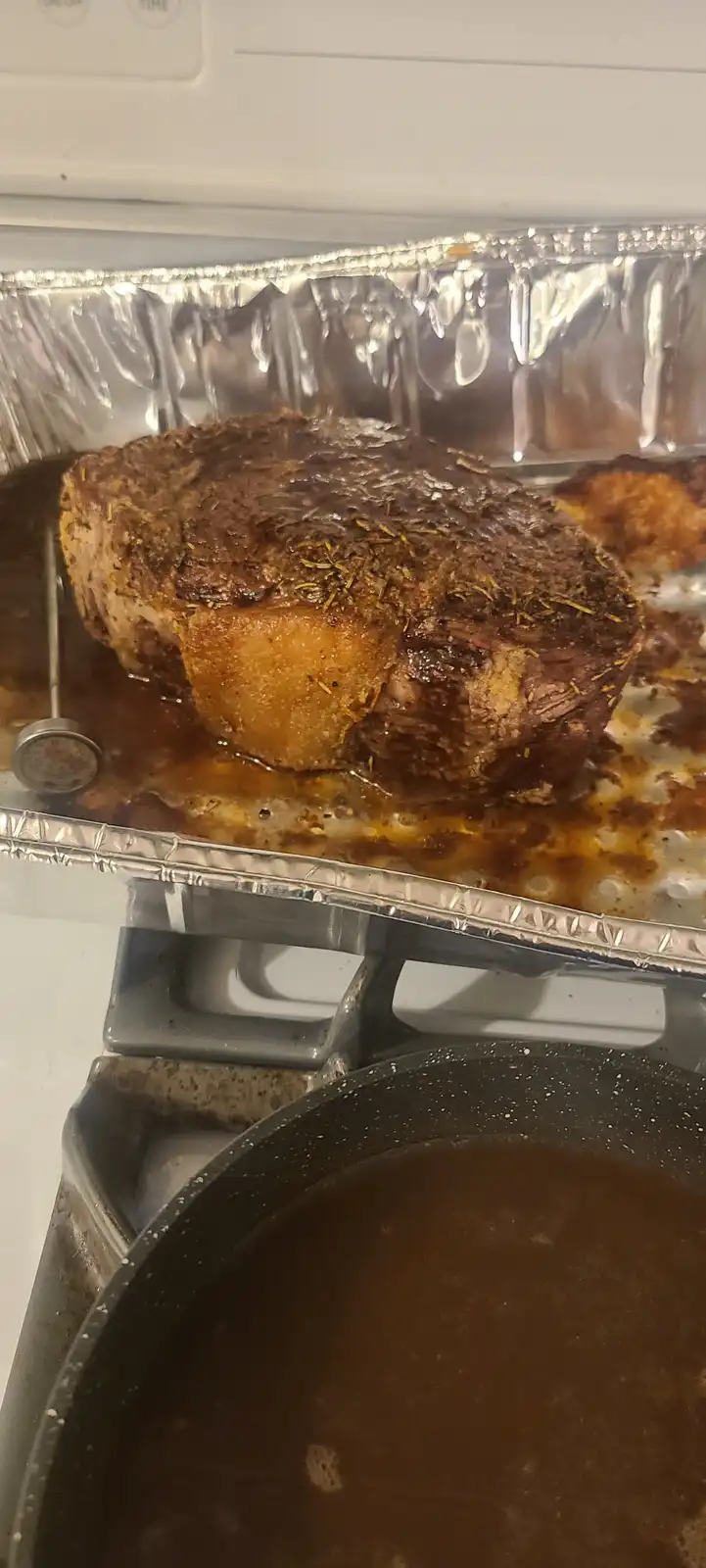A large, seared prime rib roast sits in a foil-lined roasting pan on a stovetop, with a meat thermometer inserted. Below it…
