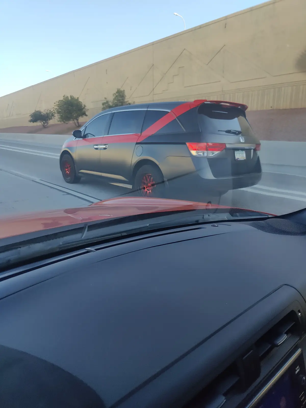 A photo taken from inside a car shows a gray Honda minivan with red accents on the roof, side stripes, and wheels, driving…