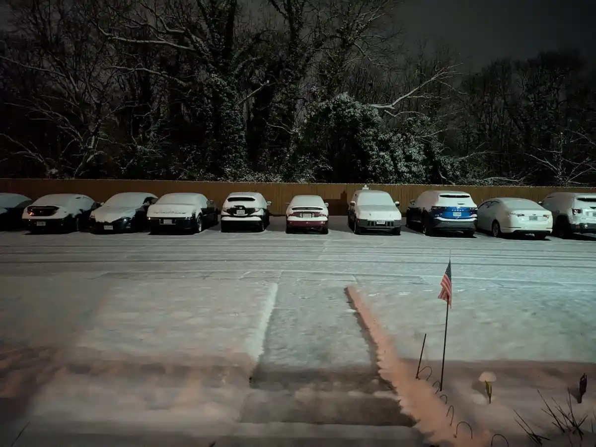 A nighttime photo of a snow-covered parking lot with several cars parked in a row, all blanketed in snow. In the foreground…