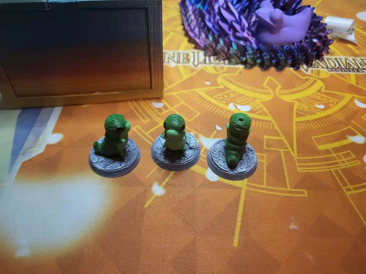 Three green, 3D-printed miniature figures of a small creature are arranged in a row on a fantasy-themed gaming mat. The…