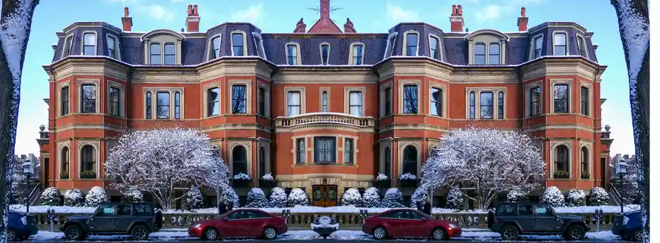 A grand, red-brick Victorian mansion with a snow-covered roof and ornate windows stands under a clear blue sky. Snow…