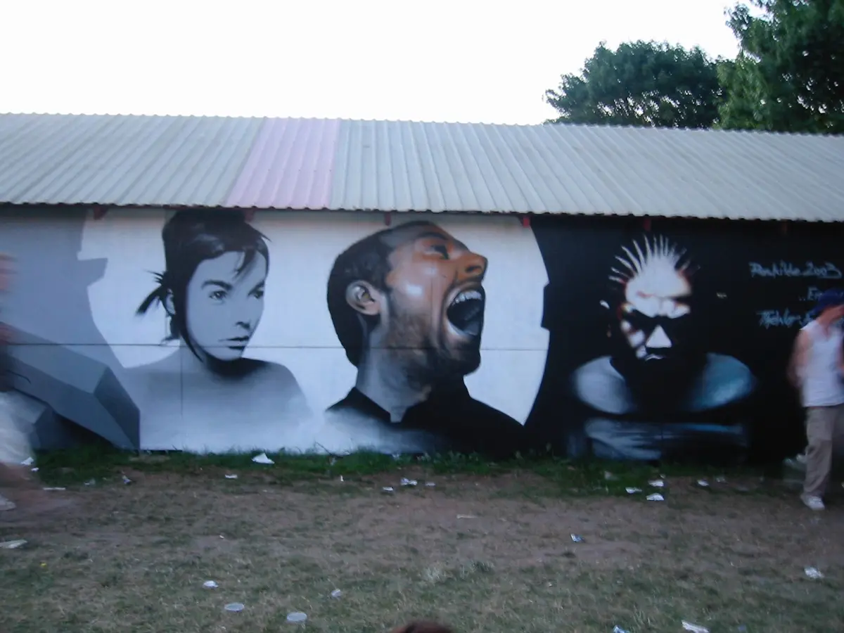 A large outdoor mural on a corrugated metal-roofed wall depicts three stylized figures: a woman with short dark hair, a man…