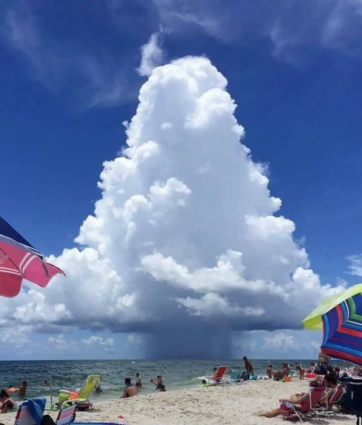 A beach scene with people relaxing under colorful umbrellas and in the water, while a massive, towering cloud shaped like a…