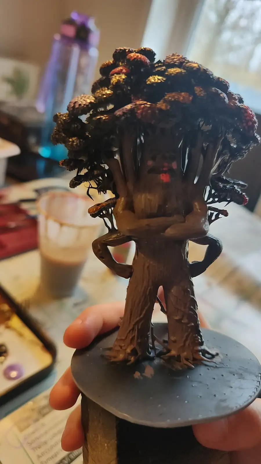 A hand holds a painted miniature figurine of a humanoid tree creature with a tree trunk body, root-like legs, and a dense…