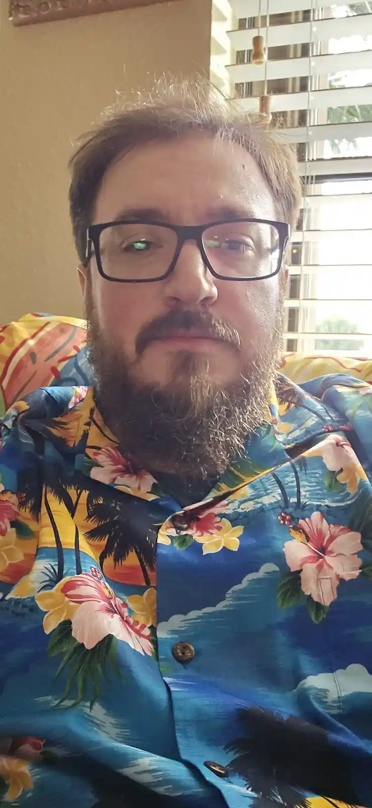 A man with glasses and a beard wears a brightly colored Hawaiian shirt featuring palm trees and flowers. He stares directly…