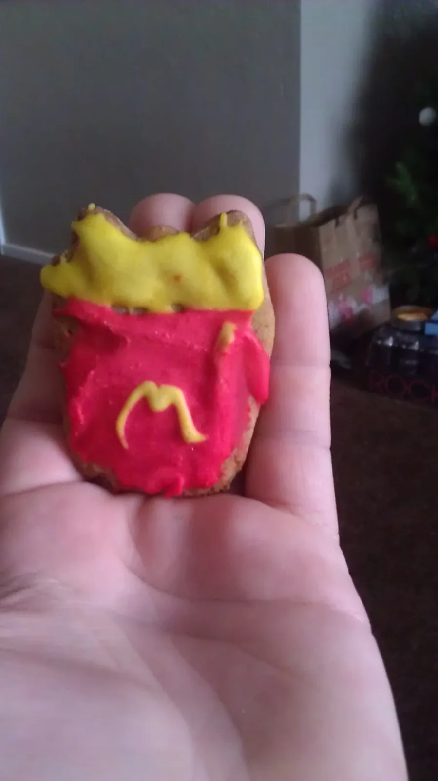 A hand holds a small, round cookie decorated with red icing to resemble a McDonald's fry container, topped with yellow icing…