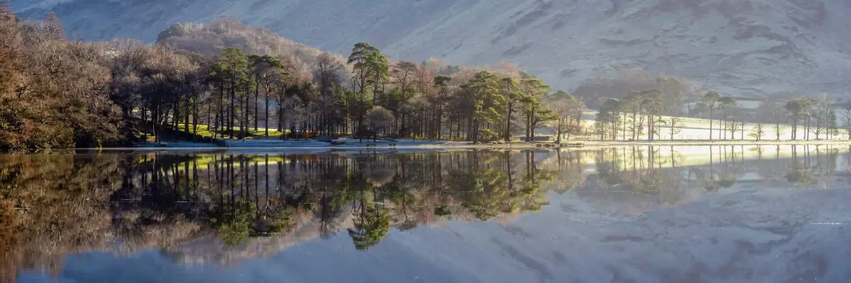 A serene winter landscape shows a calm lake reflecting snow-dusted trees and mountains under soft morning light. The…