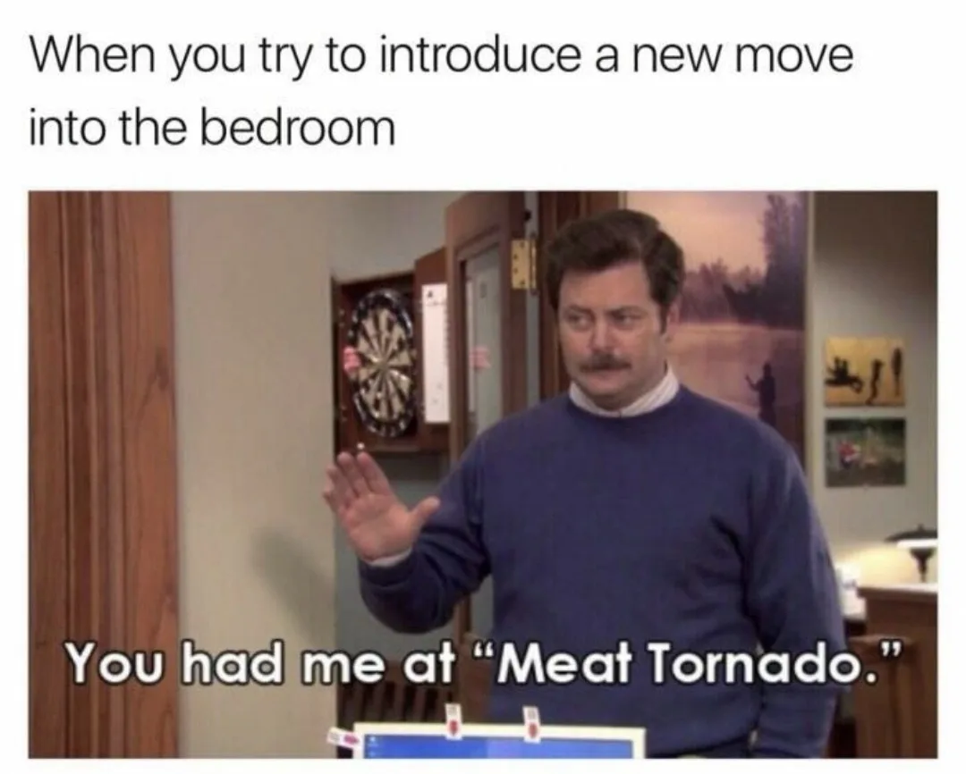 A man with a mustache wearing a blue sweater stands in a room with a dartboard on the wall behind him, gesturing with his…