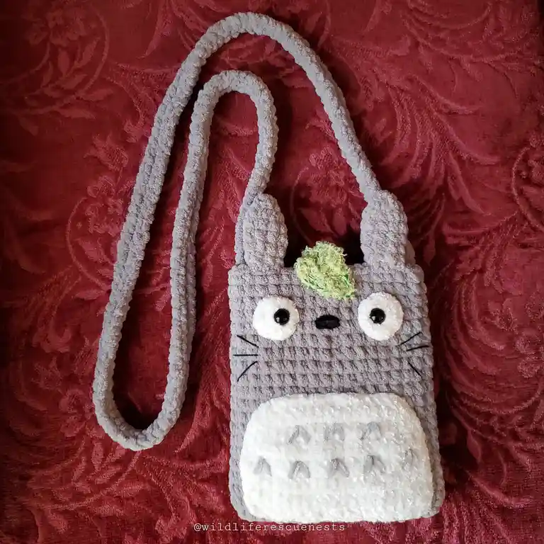 A handmade, crocheted Totoro-themed bag with large white eyes, a green moss-like tuft on its head, and a white belly with…