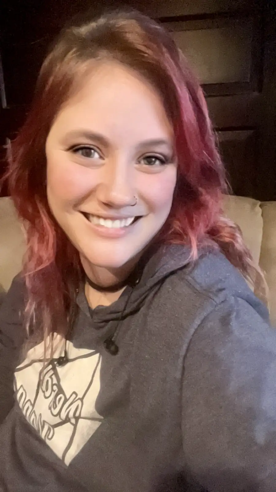 A selfie of a smiling woman with reddish-pink hair, wearing a dark hoodie with a graphic, seated on a couch. The overlaid…