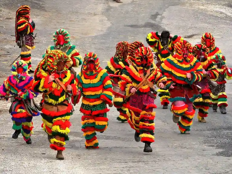 A group of people in vibrant, colorful, fringed costumes with red masks march in formation on a paved road. Their outfits…