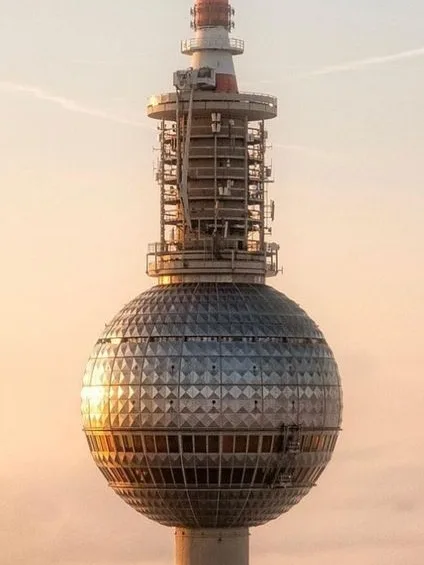 A close-up photograph of the Berlin TV Tower at sunset, showing its iconic spherical structure and antenna tower against a…