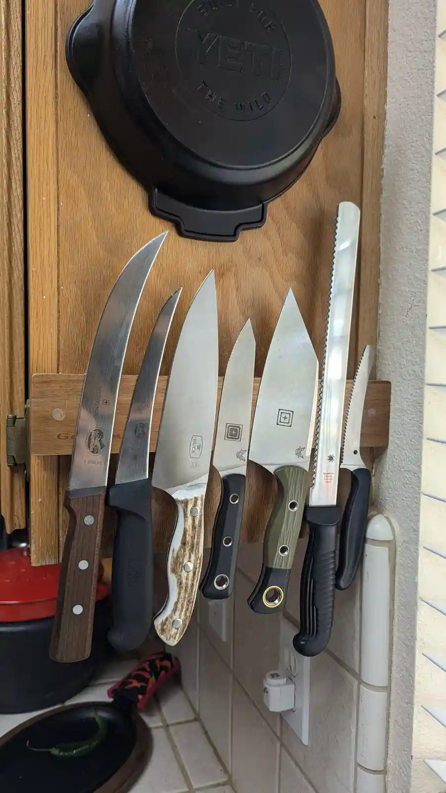 A wooden knife block mounted on a kitchen wall holds seven large, varied knives with different blade shapes and handle…
