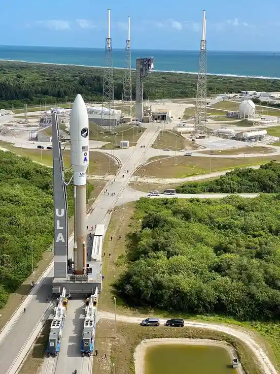 An aerial view of a large rocket, branded with ULA, standing vertically on a launch pad near an ocean, surrounded by…