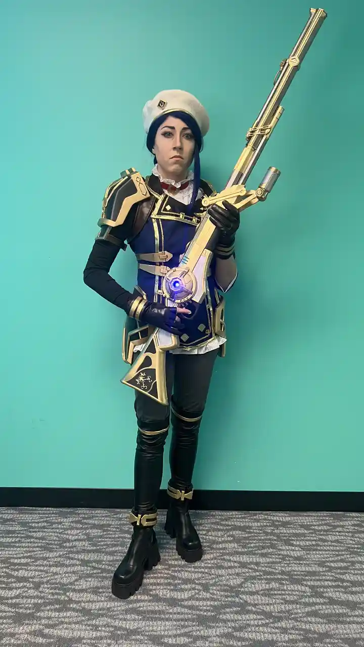 A person in a detailed cosplay of a video game character stands against a teal wall, holding a large, ornate golden and…
