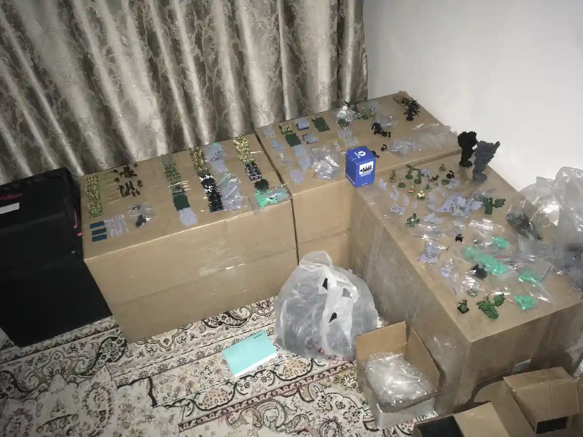 A room is filled with cardboard boxes and plastic bags overflowing with unpainted miniature figurines and terrain pieces for…