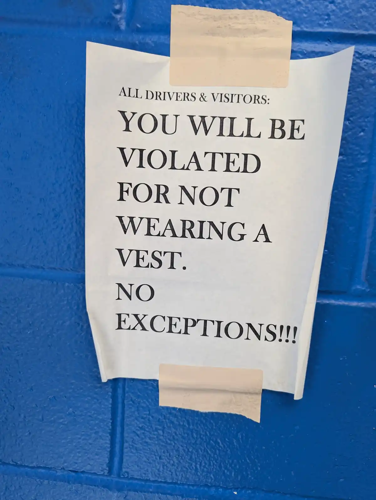A white paper sign taped to a blue brick wall with bold black text warns all drivers and visitors that they will be violated…