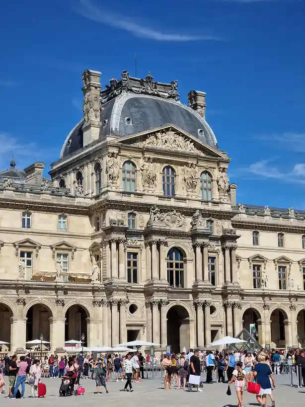 A bright, sunny photo of the ornate, classical facade of the Louvre Museum in Paris, featuring its iconic dome and detailed…