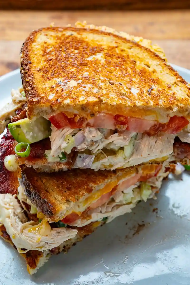 a close-up photo of a grilled sandwich cut in half, revealing layers of chicken, bacon, tomato, avocado, and melted cheese…
