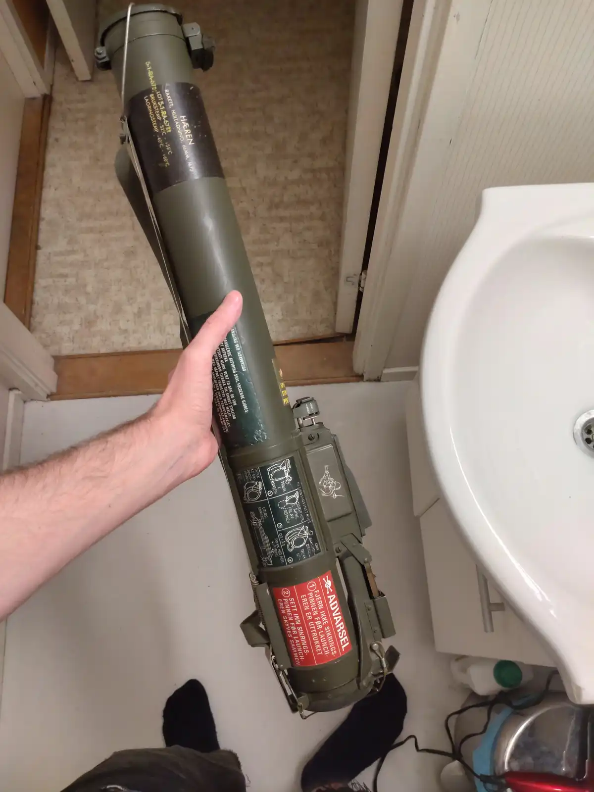 A person holds a large, olive-drab military rocket launcher in a bathroom next to a white sink, with the red warning label…