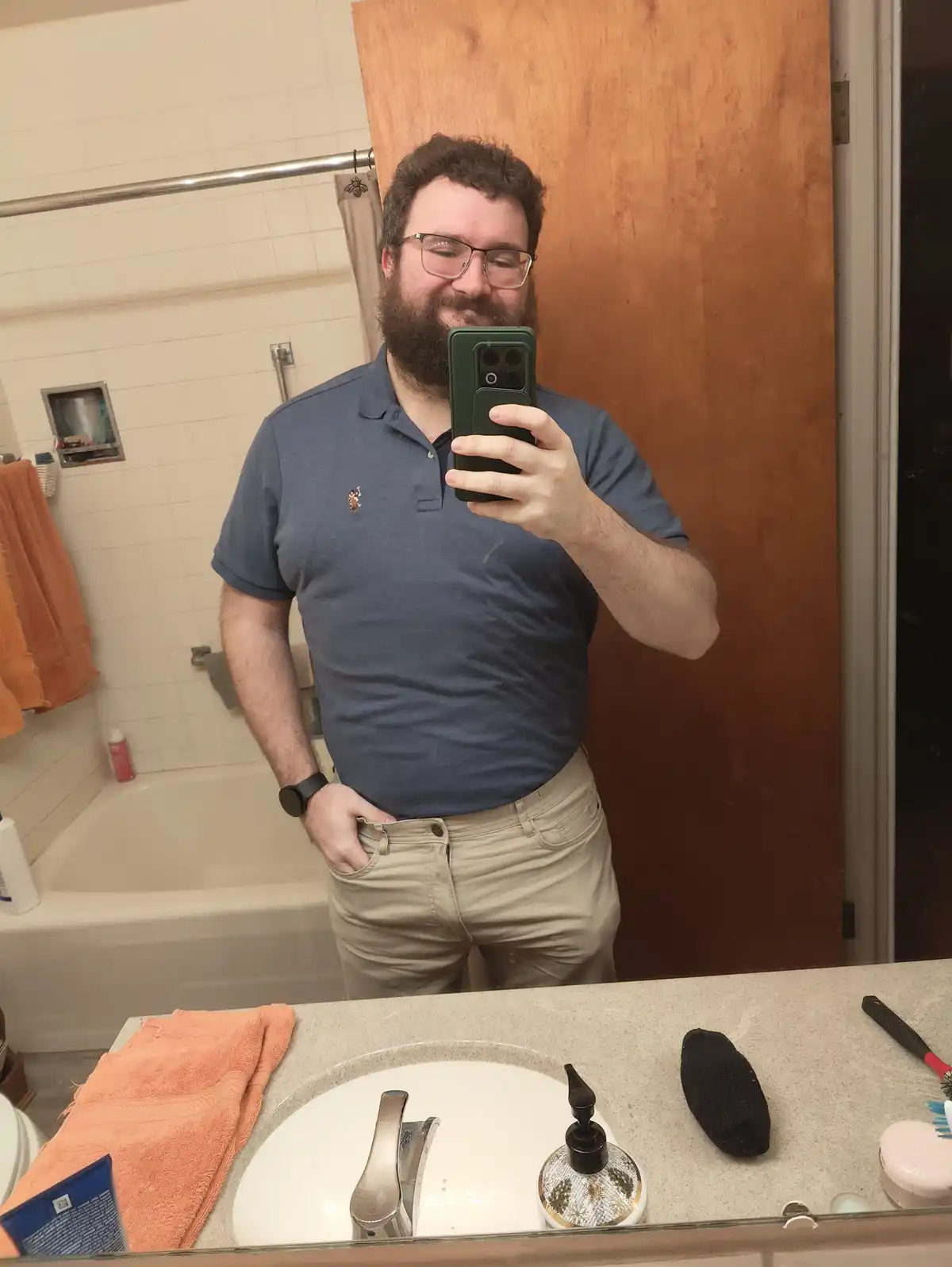 A man with a beard and glasses takes a mirror selfie in a bathroom while holding his phone. He's wearing a blue polo shirt…
