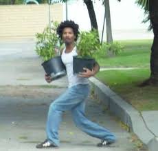 A man with curly hair, wearing a white tank top and blue jeans, stands outdoors on a sidewalk holding two potted plants, one…