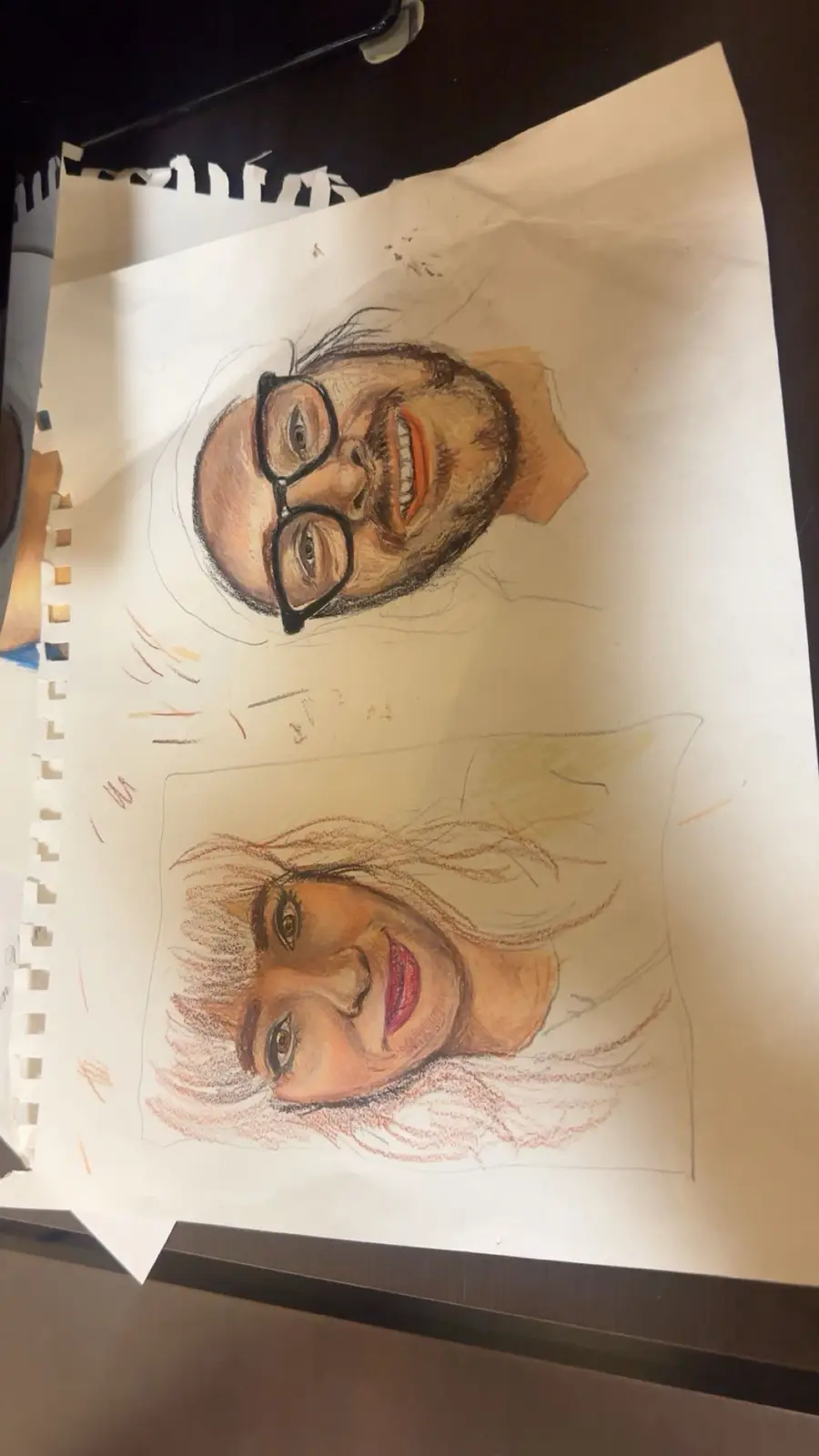 A hand-drawn sketch on paper shows two portraits: a man with glasses and a beard smiling widely, and a woman with long hair…