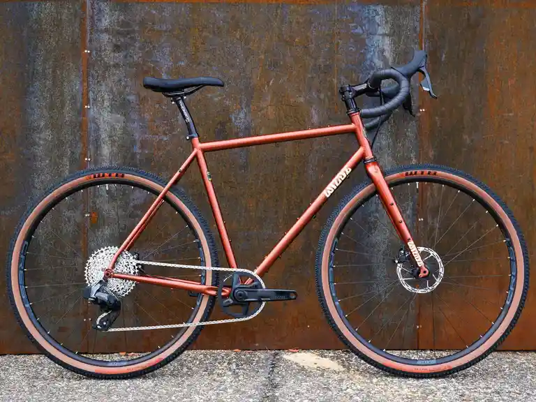 a rust-colored gravel bicycle with black handlebars and seat, standing against a rusty metal wall. the bike has tan-colored…