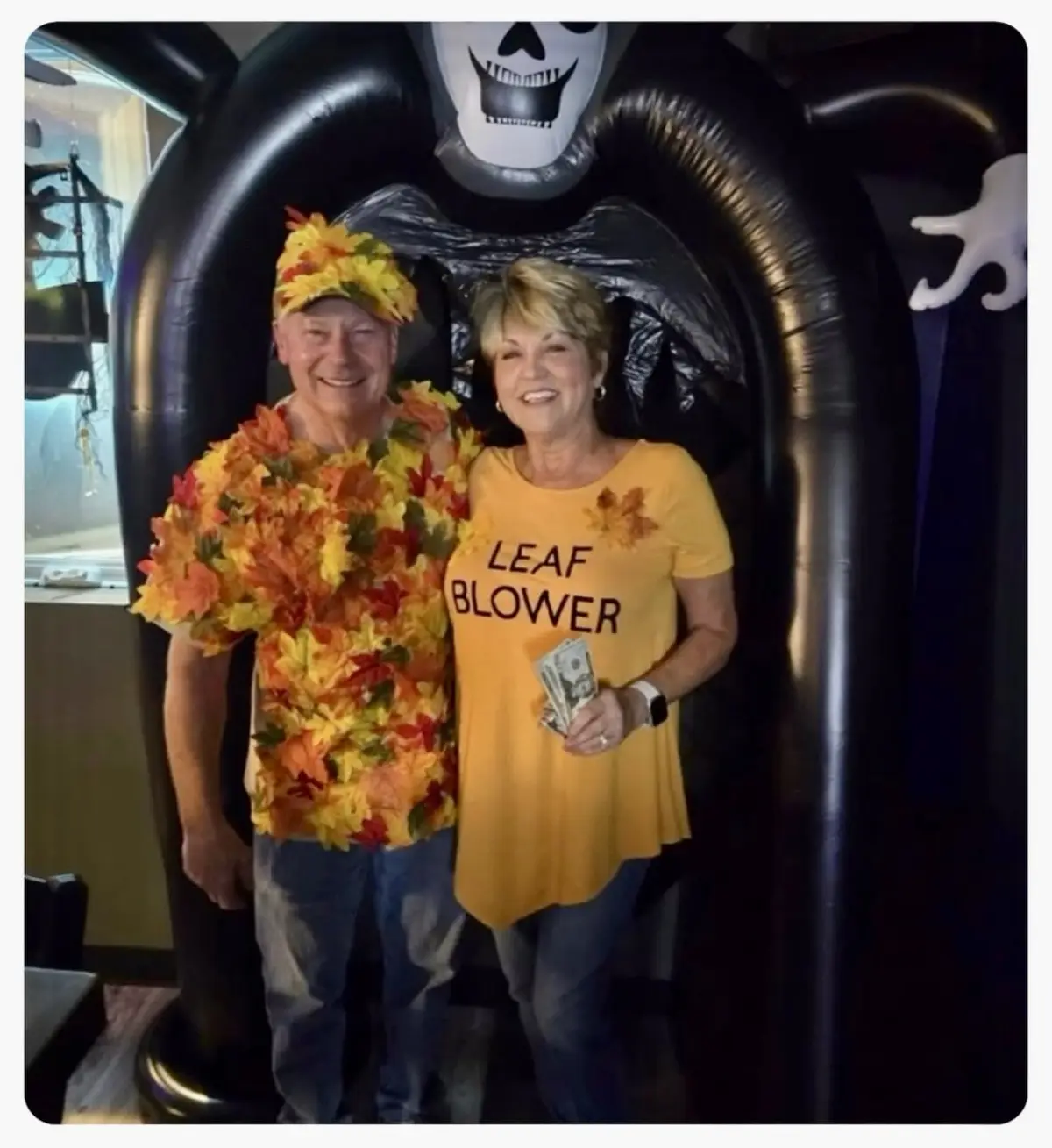 Two smiling older adults pose in front of a large inflatable Halloween skeleton arch. The man wears a costume made of autumn…