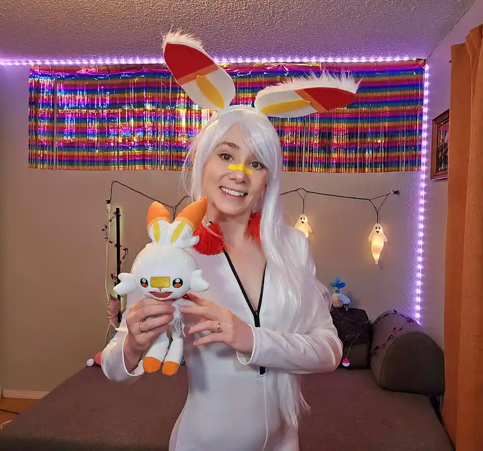 A person with white hair and large bunny ears holds a plush toy of the Pokémon 'Sobble' while wearing a yellow nose sticker…