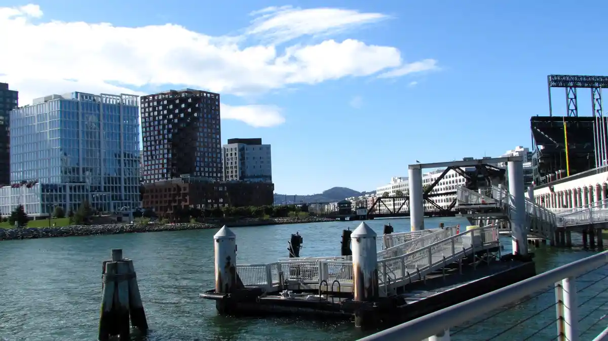 A scenic waterfront photo shows modern city buildings, a bridge, and a pier under a partly cloudy sky. Overlaid text reads…