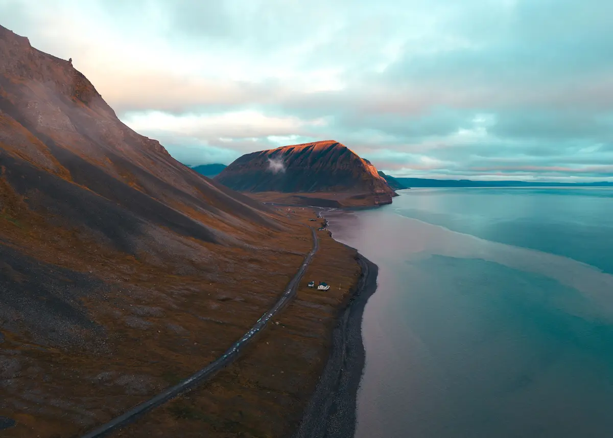 An aerial view of a remote, winding road along a rugged coastline with mountains and a calm sea under a cloudy sky. Two…