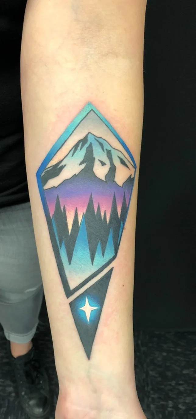 A tattoo on a person's forearm depicts a stylized mountain range within a diamond-shaped frame, featuring a gradient sky…