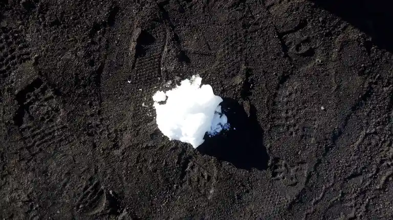 A small, bright white snowball rests on a dark, textured surface that appears to be soil or volcanic ash, casting a distinct…