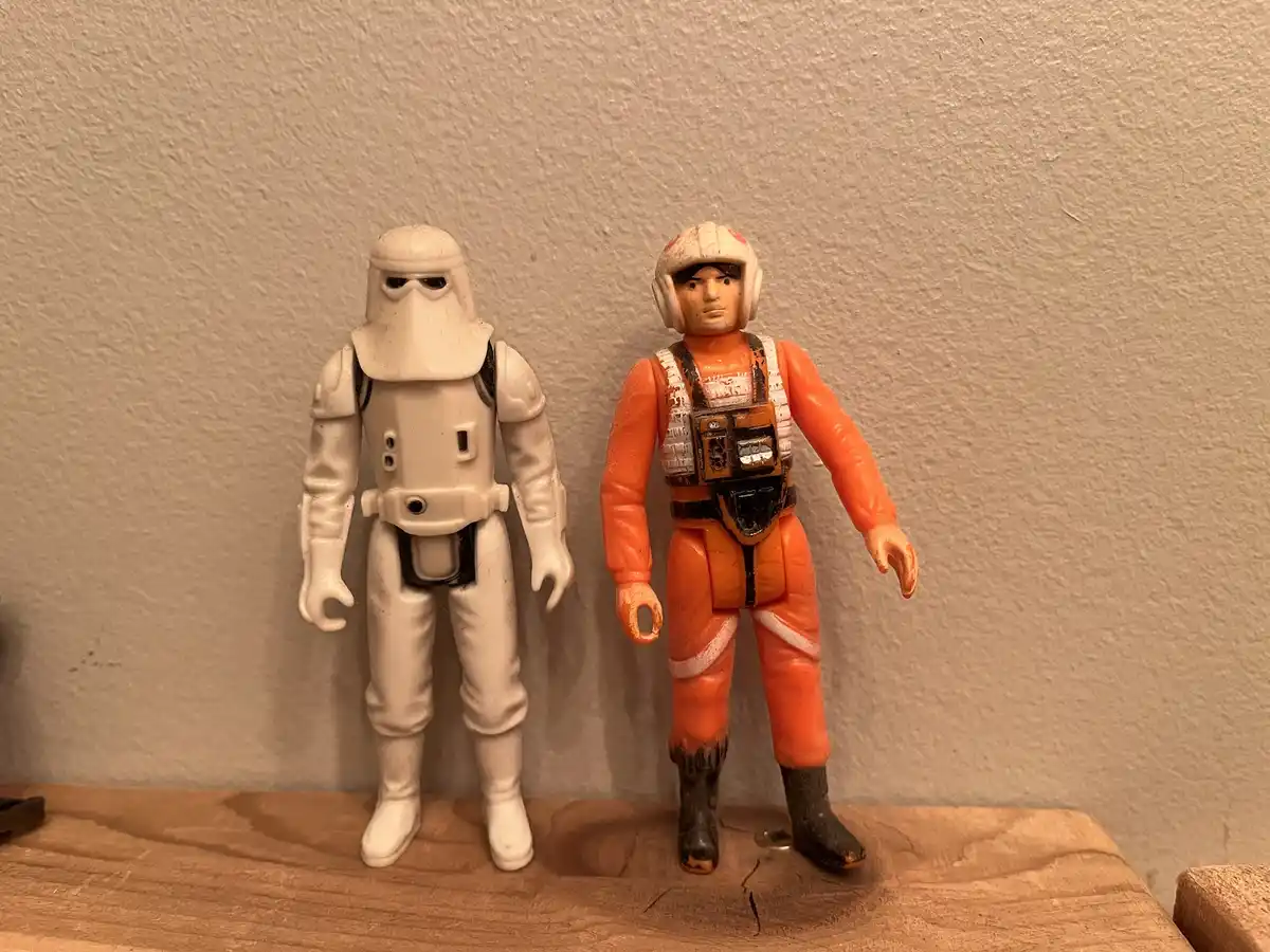 Two Star Wars action figures stand side by side on a wooden surface against a textured wall: a white-armored Stormtrooper…