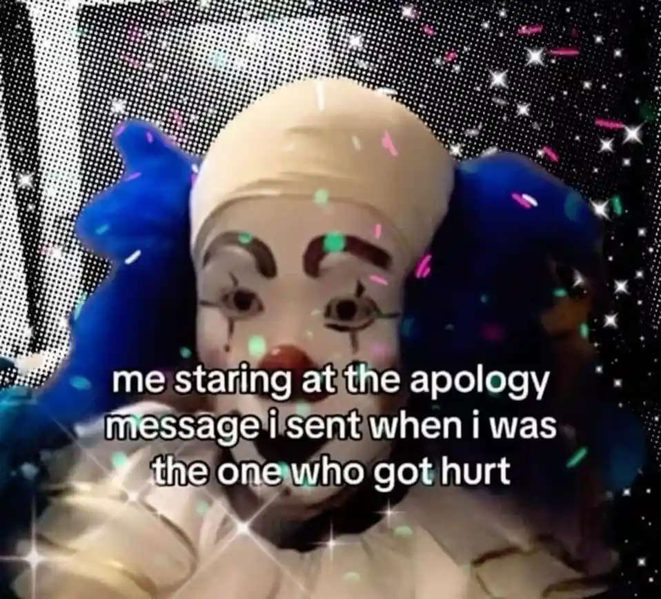 A close-up image of a clown with a white face, red nose, and blue pigtails, looking directly at the viewer with a blank…