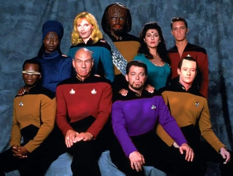 a promotional photo of the cast of 'Star Trek: The Next Generation' in their iconic uniforms, posed formally against a blue…