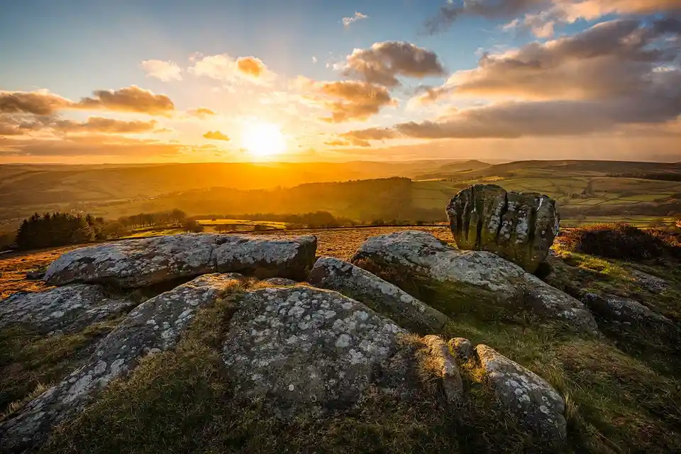 A scenic landscape photo of a sunset over rolling hills with large moss-covered rocks in the foreground. The sun is low on…