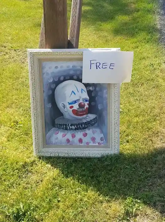 A framed painting of a creepy clown with blue eye makeup, red nose, and pink polka dots sits on grass next to a wooden post…