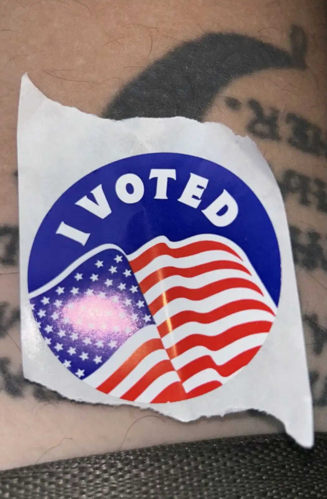 A torn 'I VOTED' sticker featuring the American flag design is stuck on a person's arm, partially covering a tattoo. The…