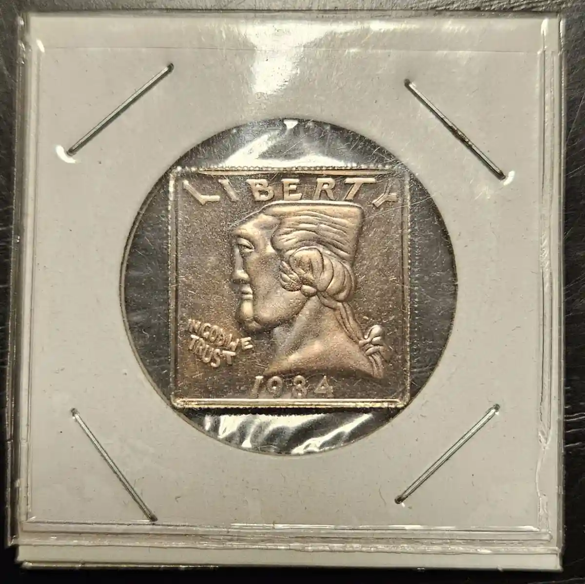 A close-up photo of a 1984 US quarter coin, encased in a clear protective holder with four metal pins securing it. The coin…