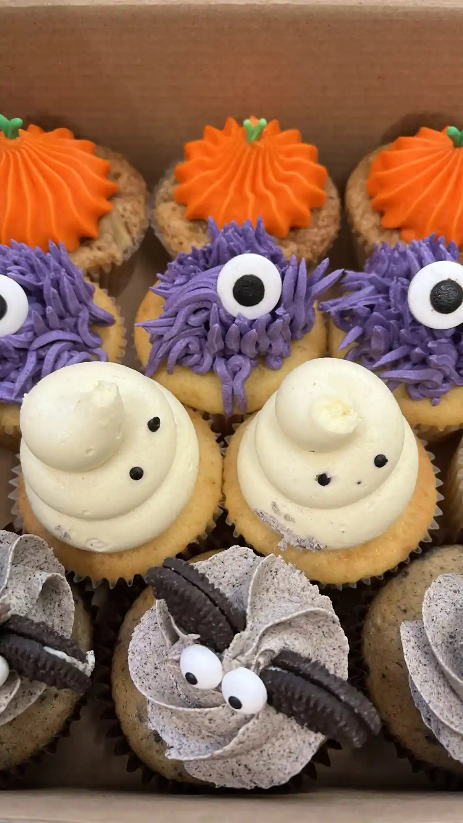 A box of Halloween-themed cupcakes decorated as pumpkins, purple one-eyed monsters, ghostly figures with small black dots…