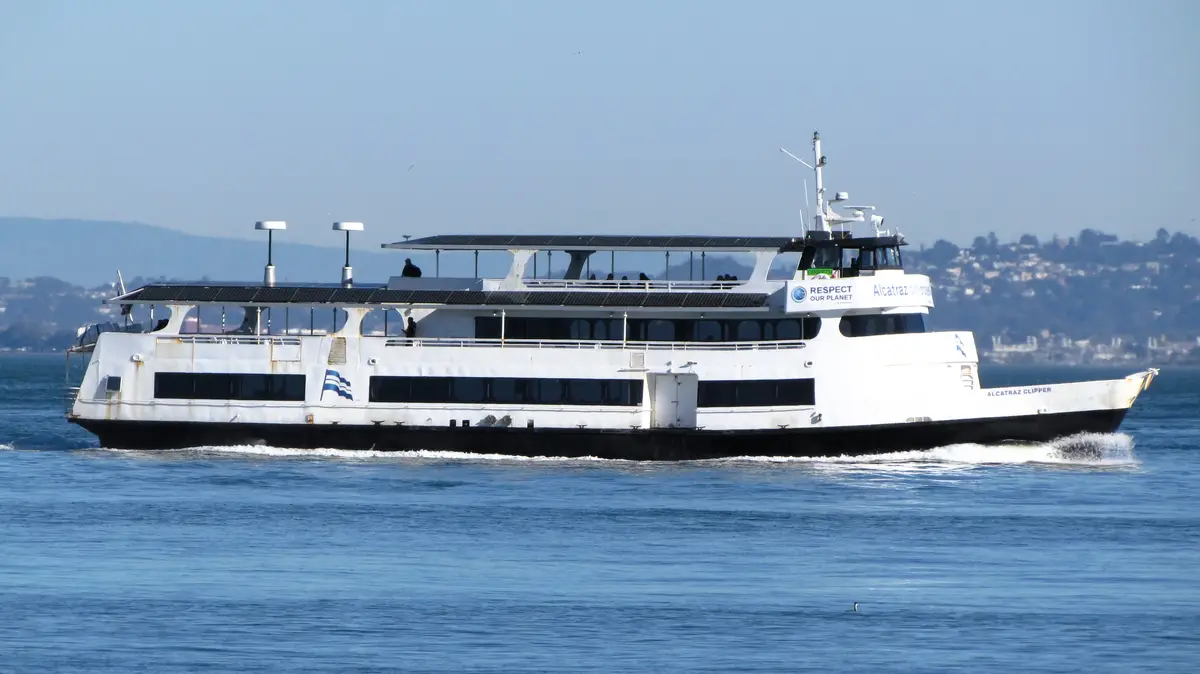 A white Alcatraz Clipper ferry with solar panels on its upper deck sails on blue water, with hills in the background. The…