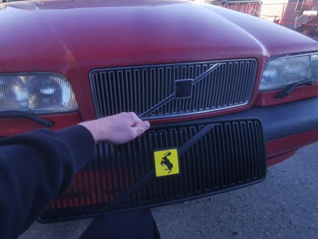 A person's hand points to the grille of a red Volvo car, which has a yellow Ferrari prancing horse logo sticker affixed to…
