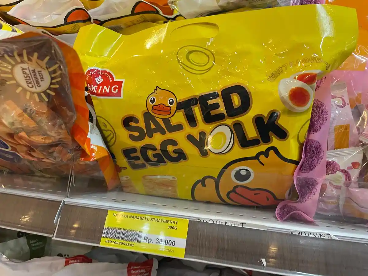 A bright yellow bag of 'SALTED EGG YOLK' snacks featuring cartoon ducks is displayed on a supermarket shelf, surrounded by…