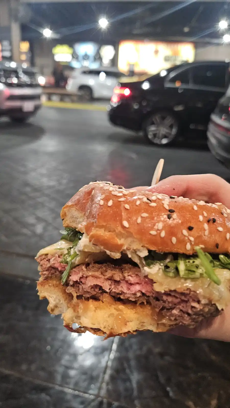 A hand holds a large, juicy burger with sesame seeds on the bun, melted cheese, and greens, in front of a blurry nighttime…