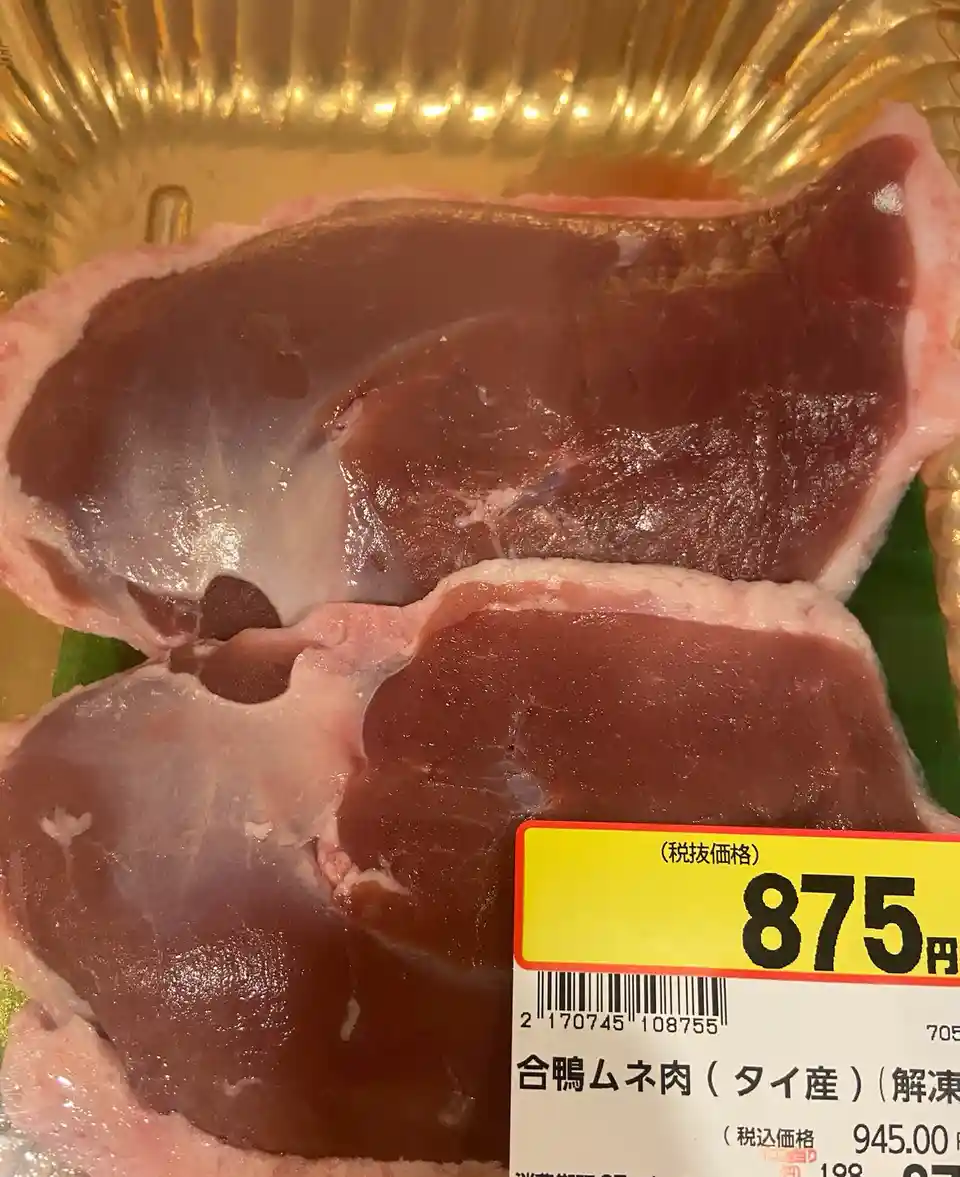 A close-up photo of two raw duck breast cuts in a gold-colored plastic tray, with a yellow price tag showing '875円'…