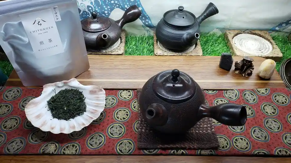 A still life of traditional Japanese tea setup featuring three dark ceramic teapots on wooden trays, a bag of 'CHIYONOEN TEA…